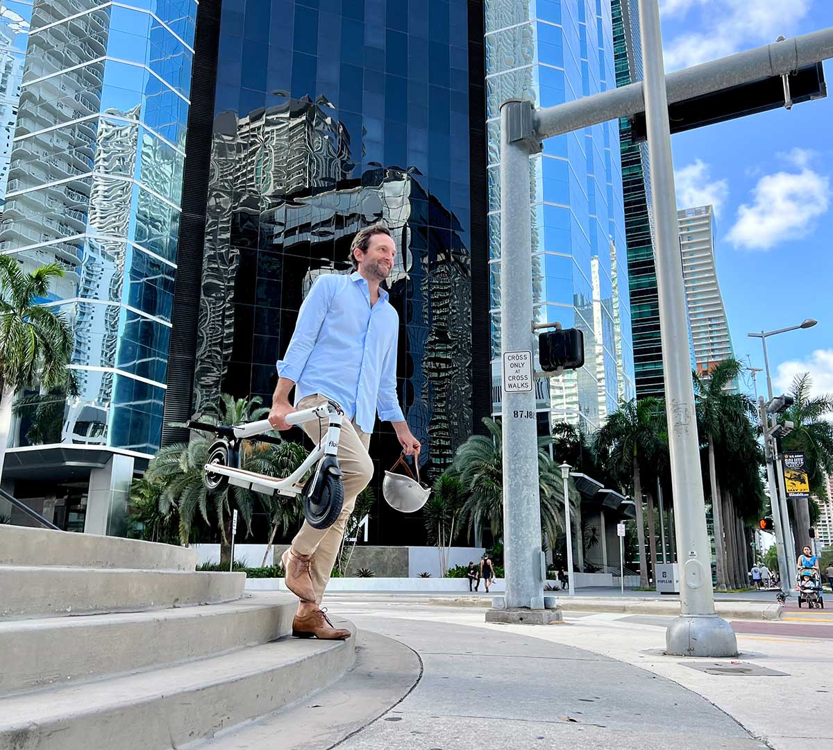 A man in casual business attire cheerfully walks with a folded electric scooter in hand across an urban landscape, demonstrating the evolution of electric scooters from trendy gadgets to practical transportation solutions in modern cities.