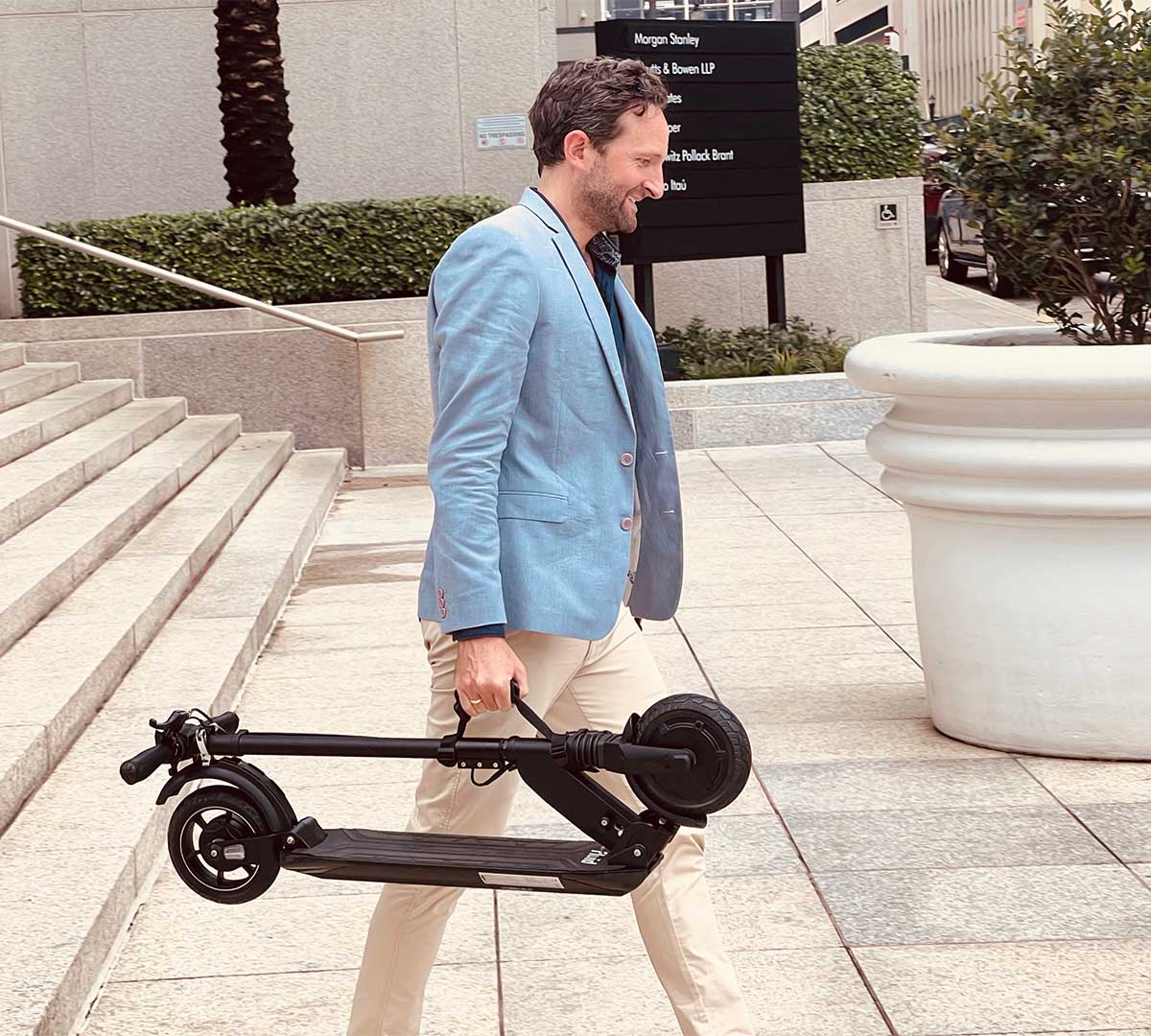 An adult male in a light blue blazer and beige trousers happily carries a small, foldable black electric scooter, ideal for city commuting, as he walks by a business complex.